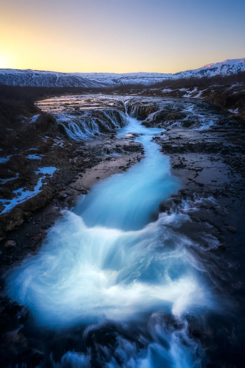 Long-exposure photo of a turquoise glacial river cascading over dark rock toward snowy hills at sunset